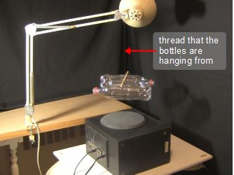 The setup showing the bottles hanging by a thread and fishing spinner from a lamp with a speaker underneath.
